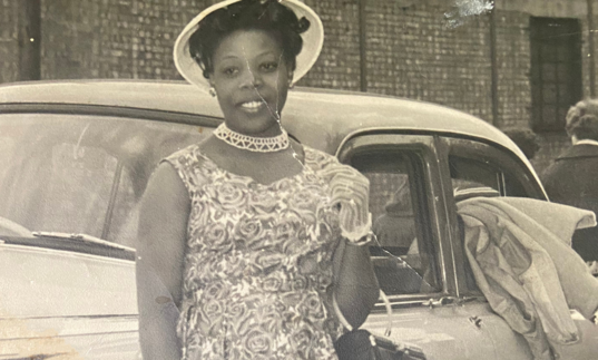 A black and white photograph of a black woman in a floral dress and white hat