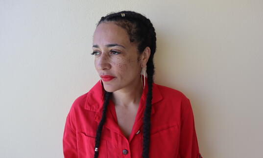 A photo of Zadie Smith wearing red lipstick and a red top, looking into the distance. 