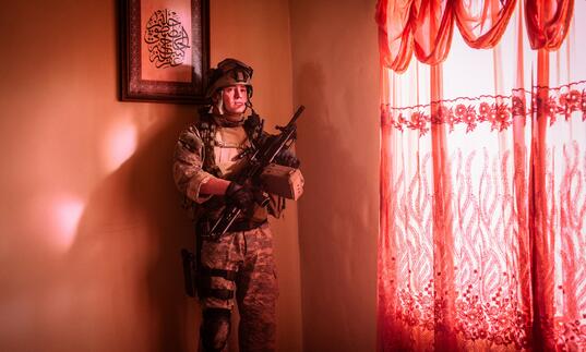 An American soldier stands in a room flooded with pink light, hiding in the corner next to the window.