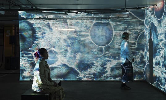 Two people sit and stand, bathed in patterned blue lights being projected onto screens