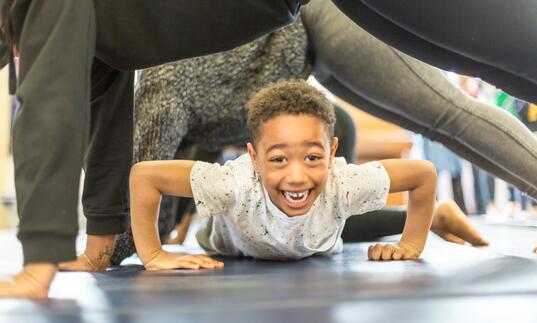 A child smiling and crawling on the ground underneath people who are planking