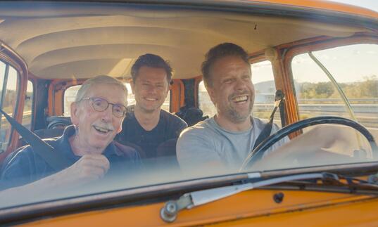 Three men sit in a yellow car, smiling