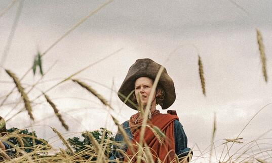 A woman wrapped in a red cloth and wearing a large leather hat stands in a field of wheat